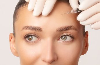 Close-up of a cosmetic professional in white gloves preparing to administer an injection into a patient's forehead. The procedure illustrates the application of dermal fillers or neuromodulators for smoothing forehead lines and wrinkles.