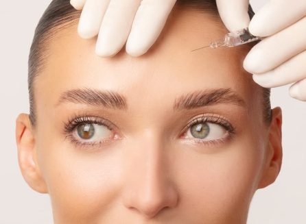 Close-up of a cosmetic professional in white gloves preparing to administer an injection into a patient's forehead. The procedure illustrates the application of dermal fillers or neuromodulators for smoothing forehead lines and wrinkles.